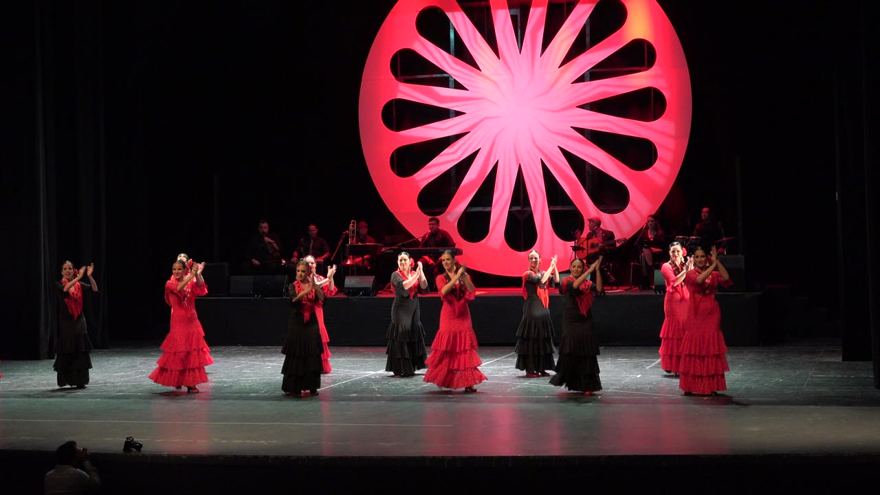 Women in red and black dresses perform across a theater stage in Cabo San Lucas, Mexico, with a red circular backdrop glowing behind them.