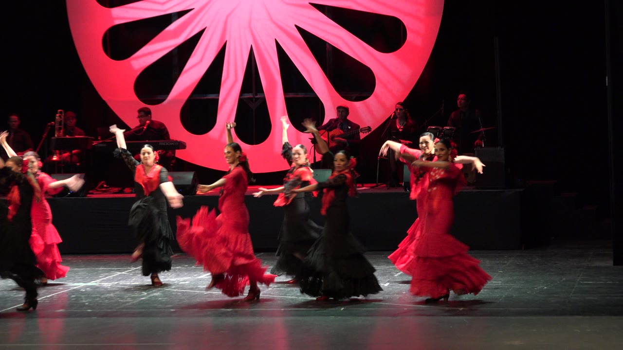 Women in red and black dresses dance on a stage with a red backdrop and musicians behind them.