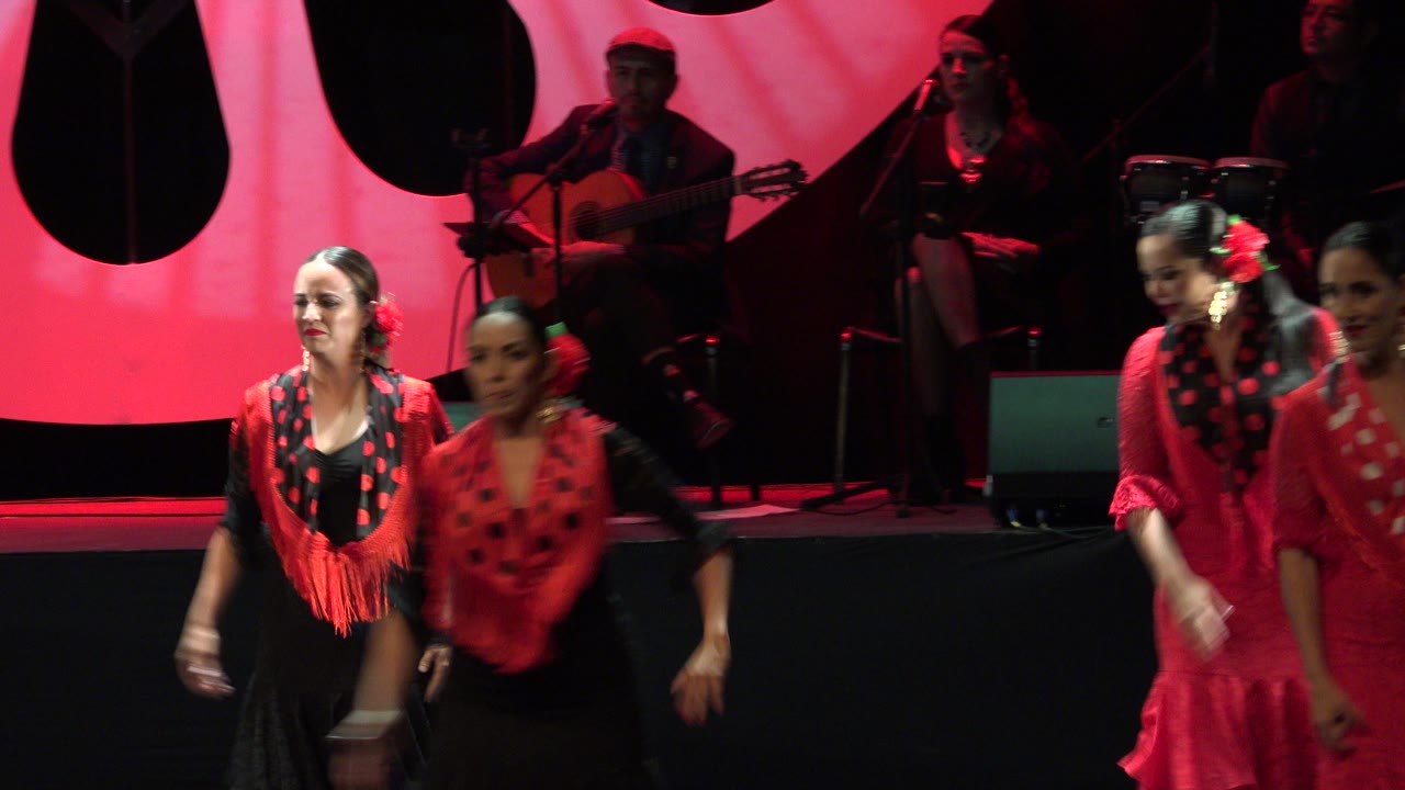 Dancers in red and black perform on a stage with musicians behind them.