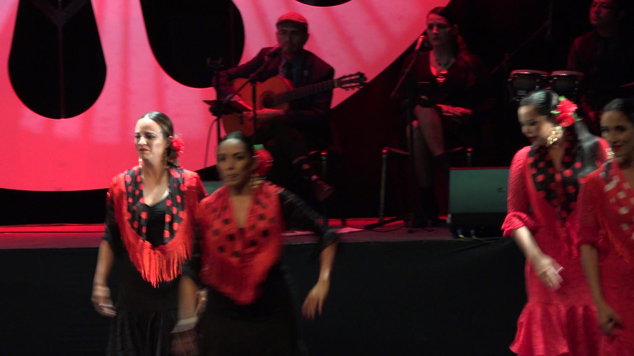 One second in, several women in red and black costumes move through the foreground as the red backdrop and seated band remain visible behind them in Cabo San Lucas.