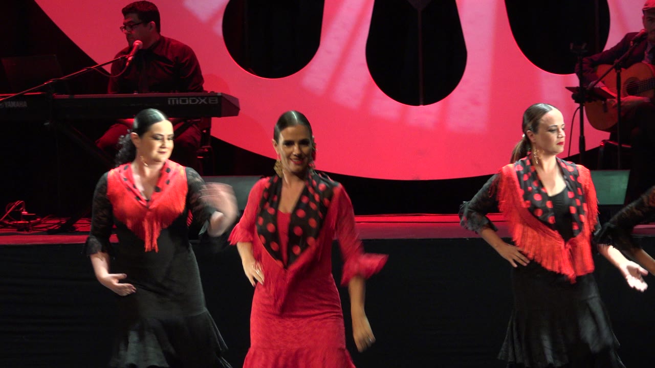 Three women in coordinated red and black flamenco-style stage costumes appear across the frame during the Cabo San Lucas performance.