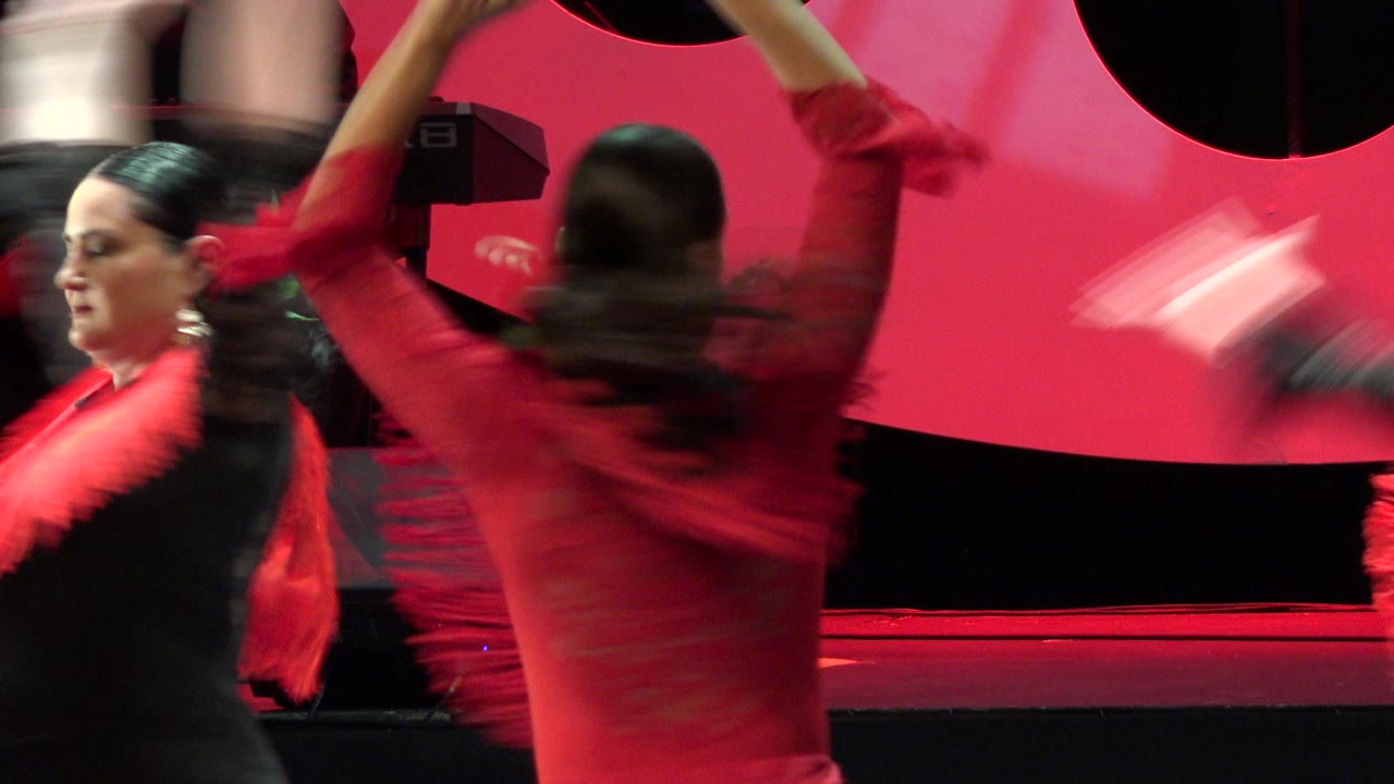 Two performers continue the indoor Cabo San Lucas dance, with one woman's profile visible and another dancer's raised arms blurred in front of the red background.