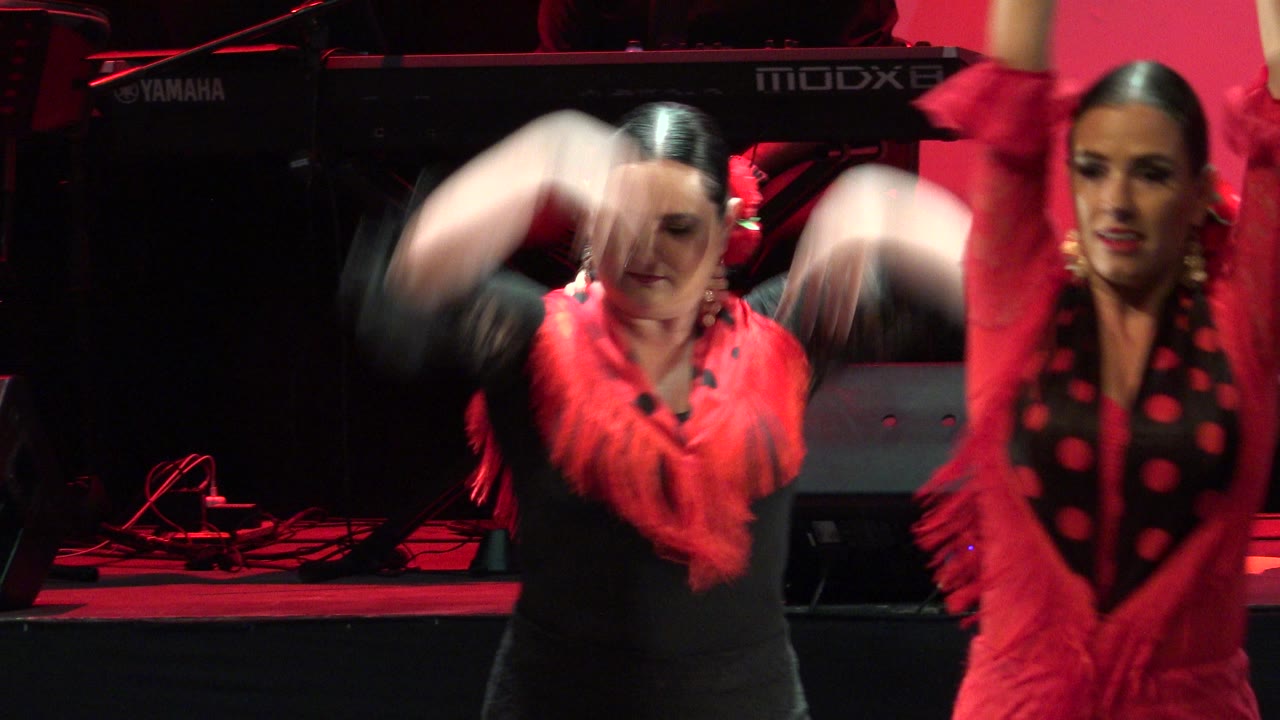 Two women perform on a red-lit stage in Cabo San Lucas, Mexico, with blurred raised arms, red shawls, black dresses, and a Yamaha keyboard behind them.