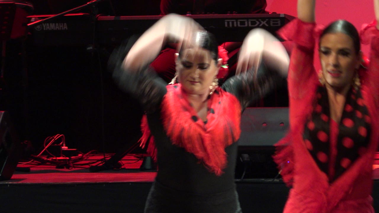 The dancers continue in Cabo San Lucas, Mexico, with one woman centered under red light while another in a red dress and black dotted shawl moves at the right edge.