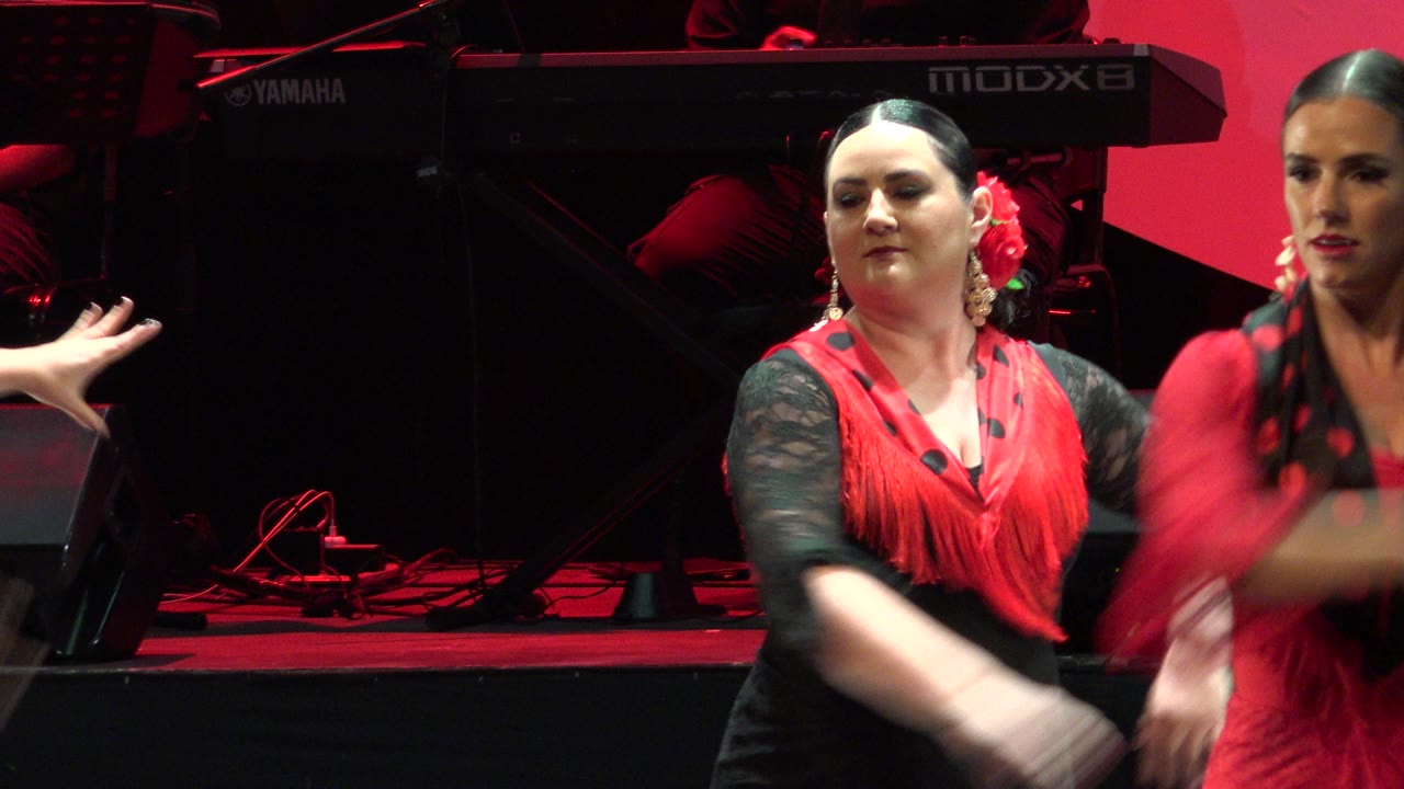A woman in black and red dances beside another performer at the edge of frame.