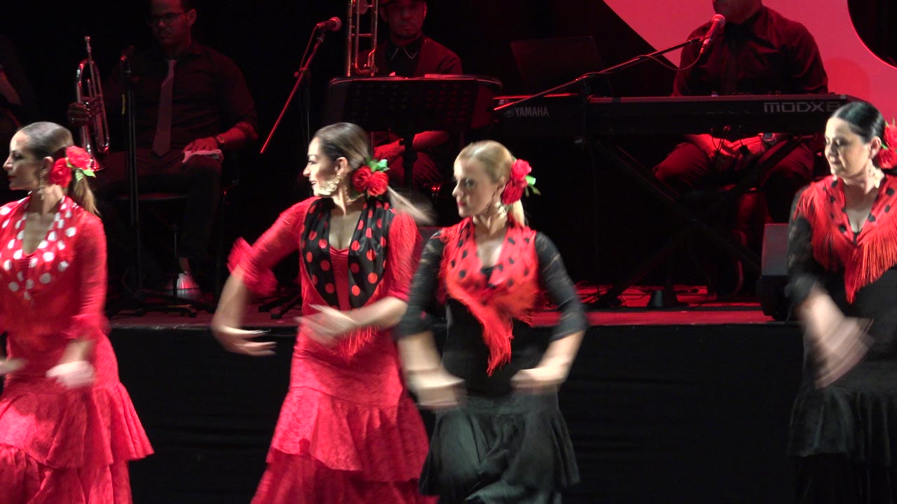 Adult women in red and black costumes perform a choreographed stage routine in Cabo San Lucas, Mexico, with musicians dimly visible behind them.