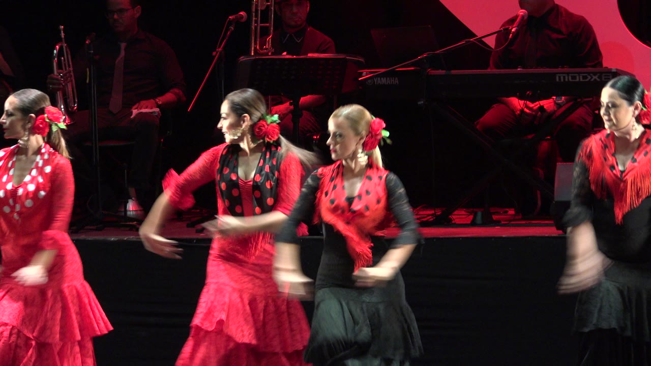 Women in red and black stage outfits continue the Cabo San Lucas dance routine while their arms blur from fast synchronized movement.