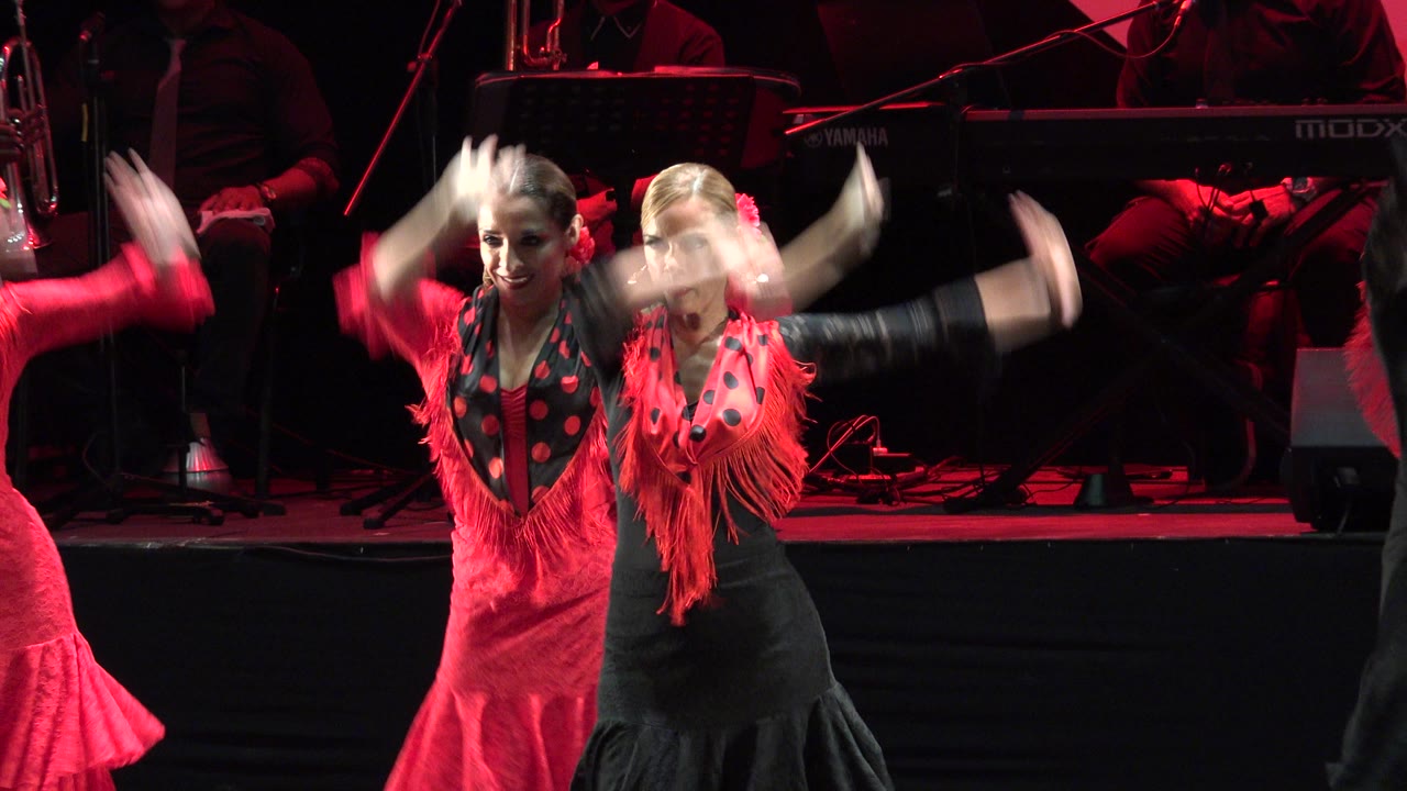 Two dancers raise their arms in front of a dark stage band.