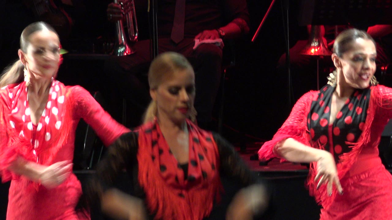At 0:00 in Cabo San Lucas, two women in red and black stage costumes are seen mid-routine, with one blurred arm crossing the foreground and dark instruments behind them.