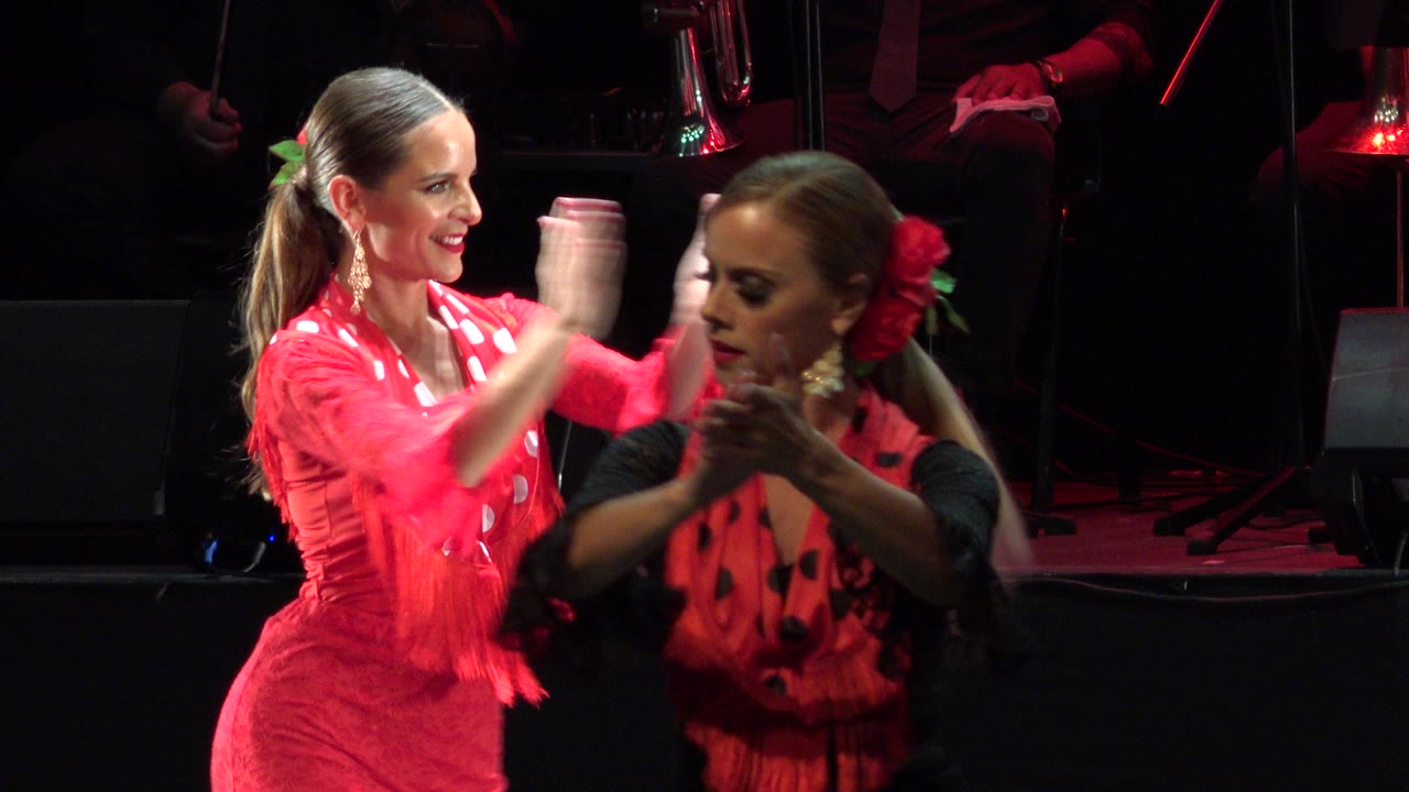 A woman in a pink fringed dress smiles and claps on a Cabo San Lucas stage while a second performer in red and black passes across the foreground.