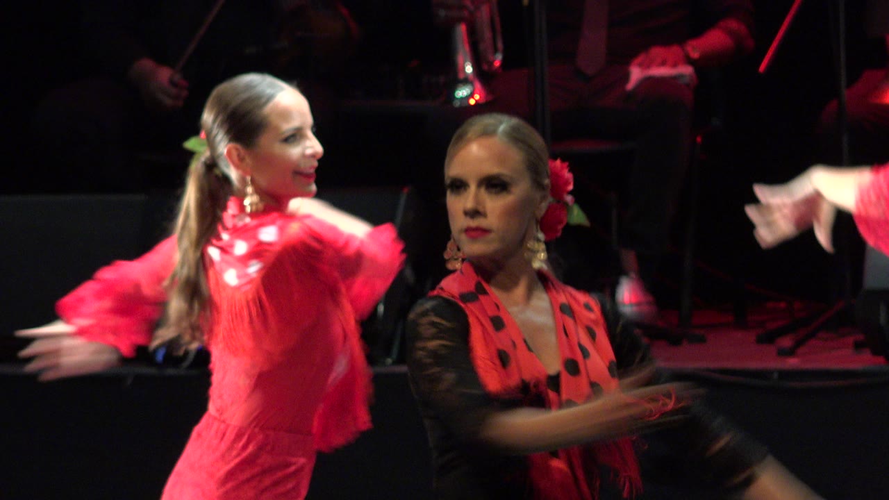 The dancers continue the Cabo San Lucas stage performance, with the woman in red turning beside the woman in black sleeves as blurred hands cross the frame.