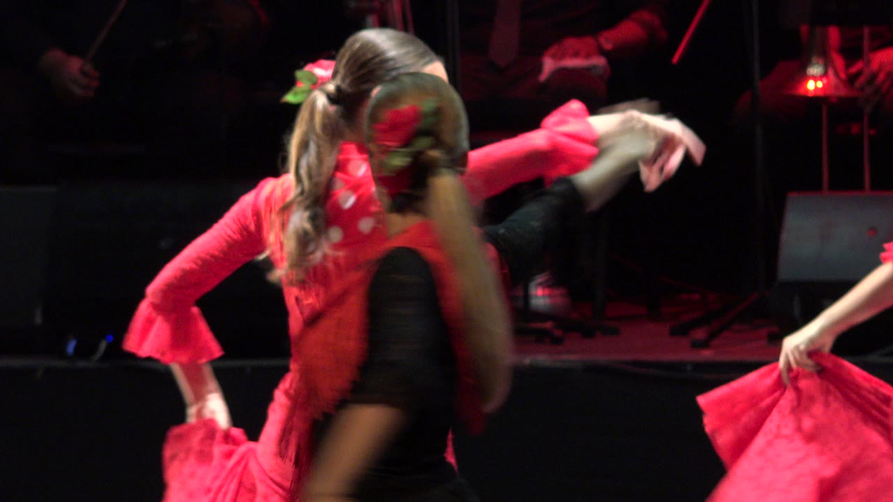 A close stage view in Cabo San Lucas, Mexico shows overlapping women dancers, red sleeves, a black dress, and a ruffled red skirt entering from the right.