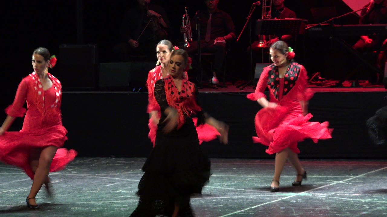 Dancers in red and black dresses perform on a dark stage.