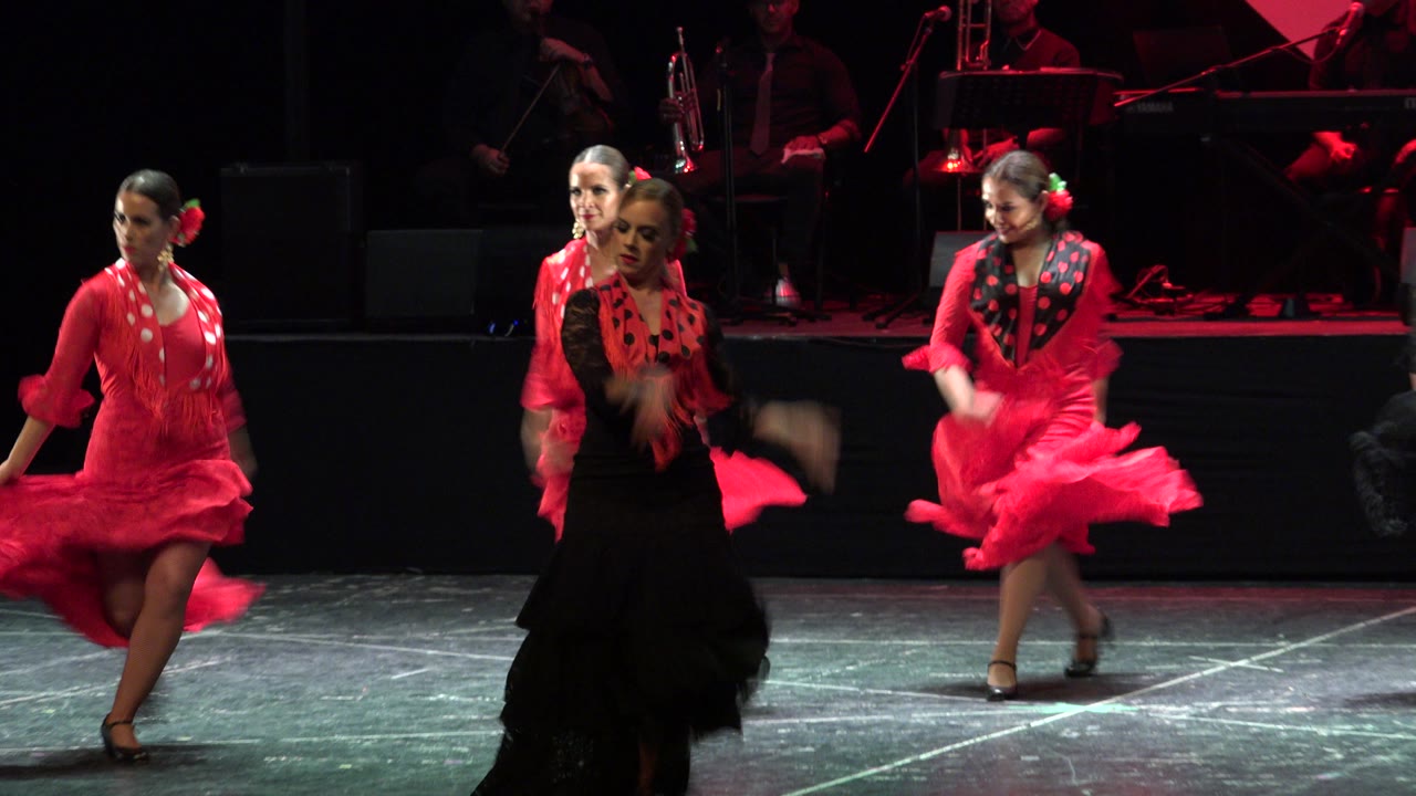 In Cabo San Lucas, Mexico, three dancers in red and black dresses cross the stage, with a red skirt flaring and the band dimly lit behind them.