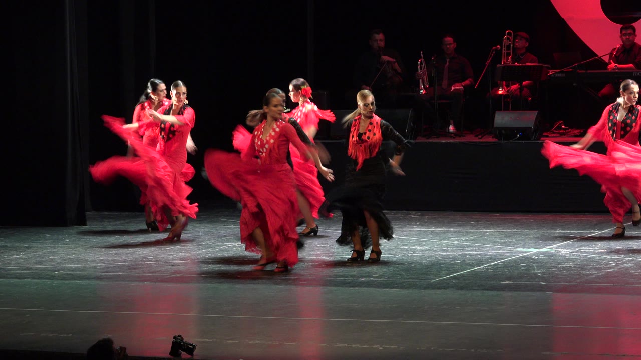 A dancer in black steps between women in red dresses during the Cabo San Lucas stage performance, with the band dimly visible behind them.