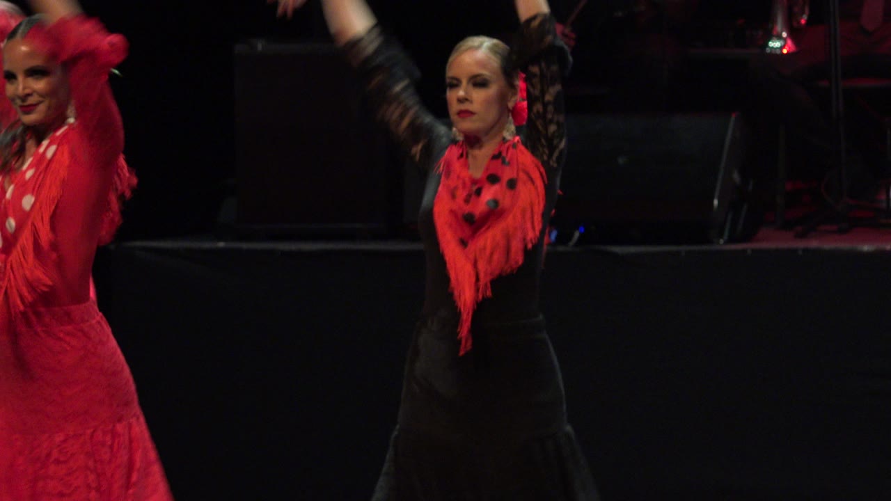 Flamenco dancers on a dark stage, one in black with a red shawl and another in red at the edge.