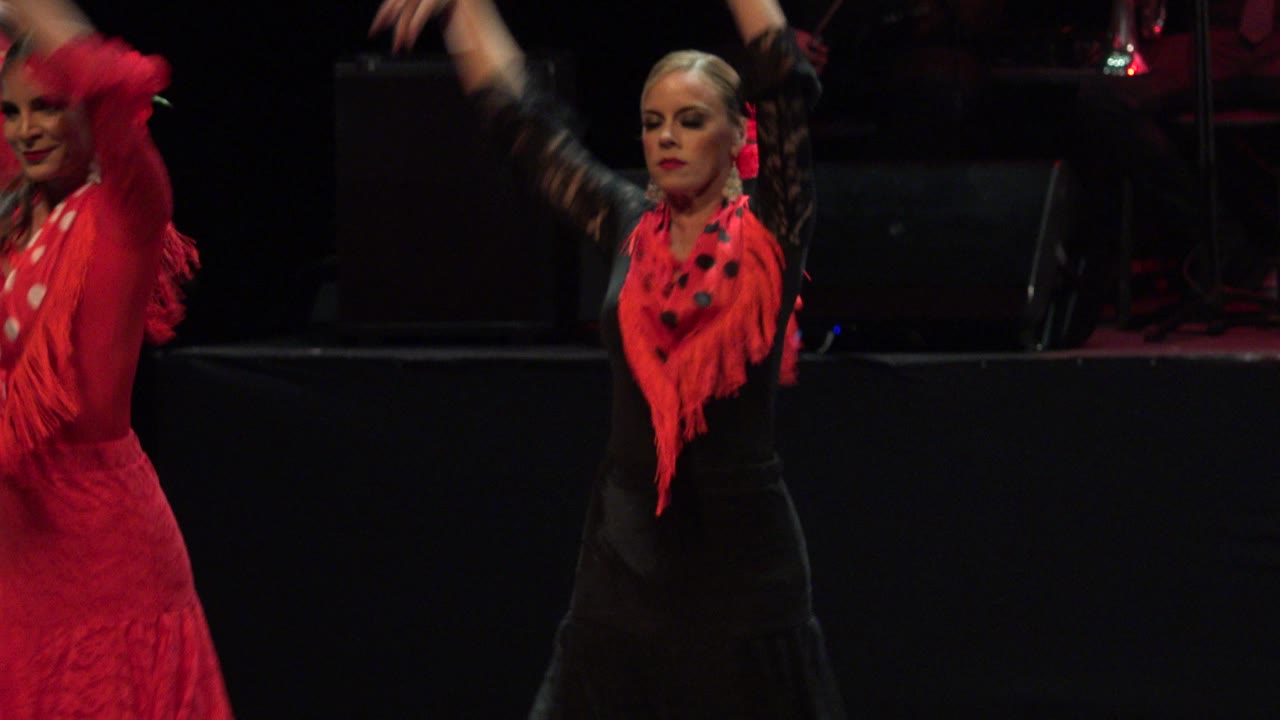 The lead dancer faces forward with both arms lifted, wearing a black dress and red shawl under low red stage lighting in Cabo San Lucas, Mexico.