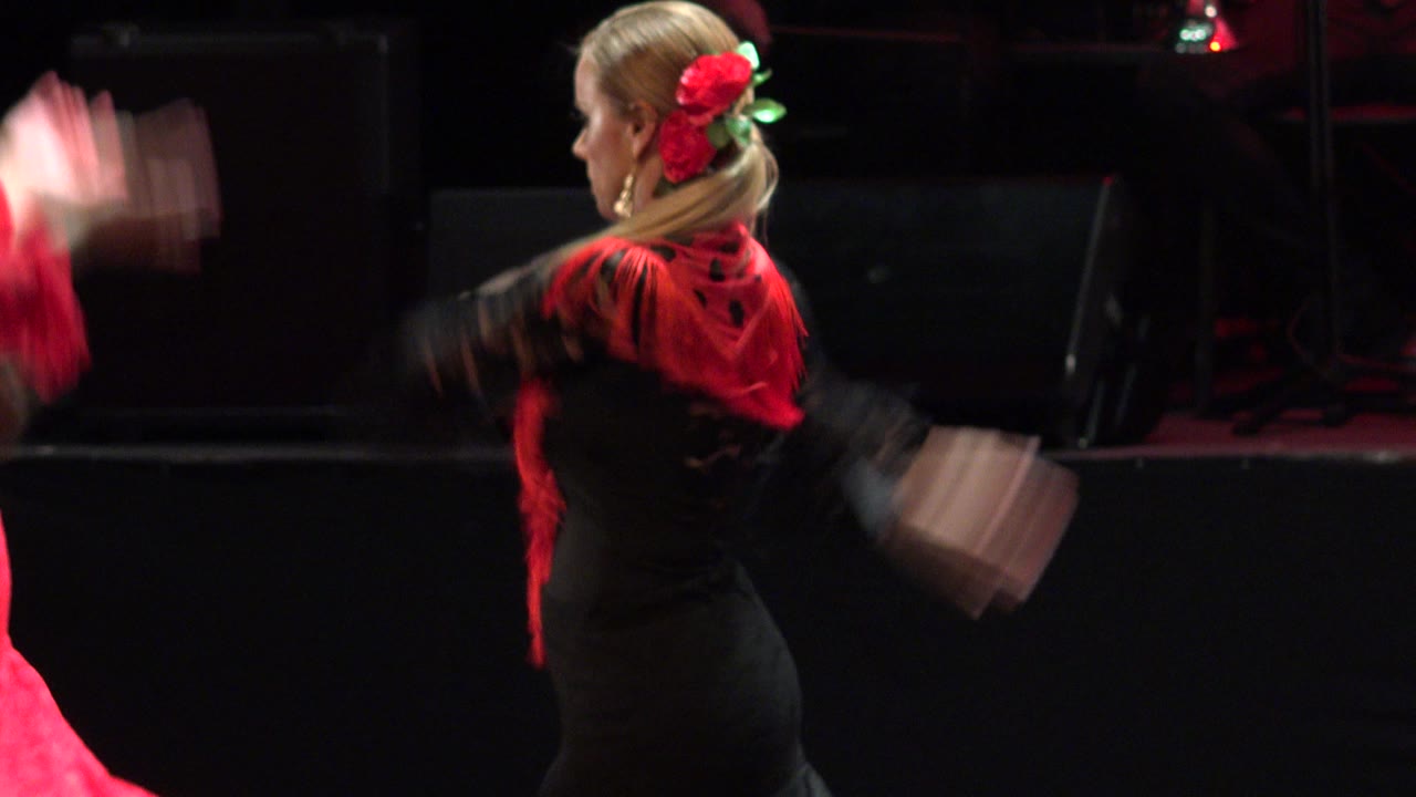 The dancer remains in side view on the Cabo San Lucas stage, with motion-blurred arms and a red costume partially entering the left edge.