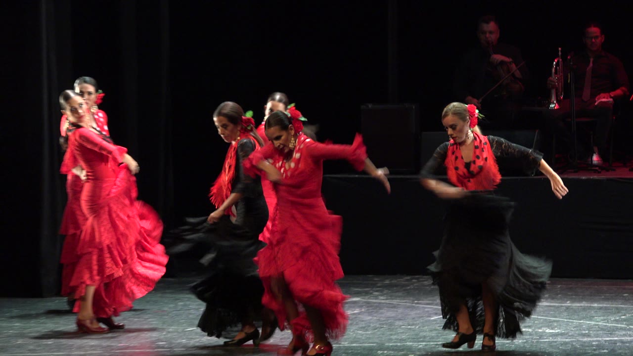 Female dancers in red and black ruffled dresses perform on a dark theater stage in Cabo San Lucas, Mexico, with musicians dimly visible at the back.