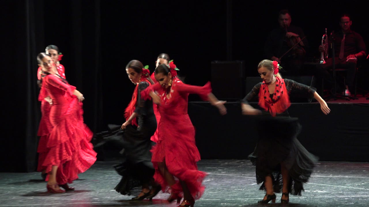 The dancers continue the stage routine in Cabo San Lucas, Mexico, with red skirts blurred by movement and background musicians in low red light.