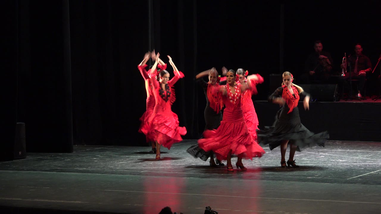 Wide view of the Cabo San Lucas performance with dancers clustered near center stage, arms raised and red skirts caught in motion.