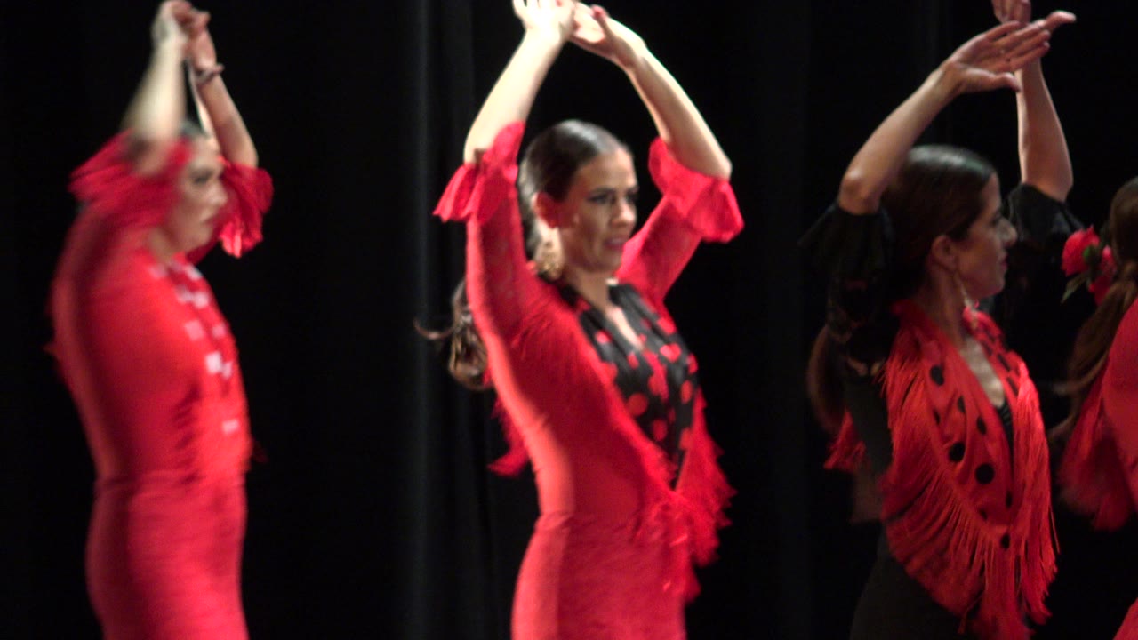Adult women in Cabo San Lucas dance on a dark stage, with one red-dressed performer raising her arms while another wears a black-and-red polka dot shawl.