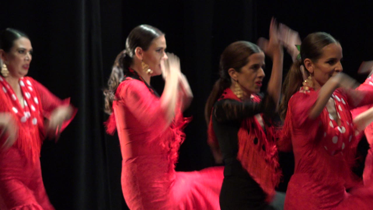 Several adult women in coordinated red and black dance outfits move across the dark Cabo San Lucas stage, with one performer turned away and others clapping in profile.