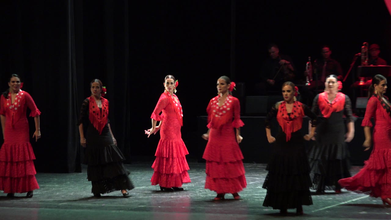 Several women in red and black ruffled dresses perform on a theater stage in Cabo San Lucas, Mexico, with musicians dimly visible in the background.