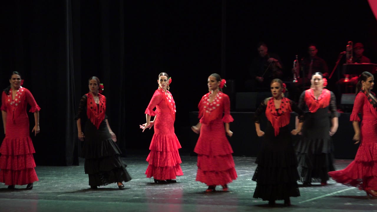The dancers hold positions across the lit stage in Cabo San Lucas, Mexico, with red costumes catching the spotlight against a black curtain.
