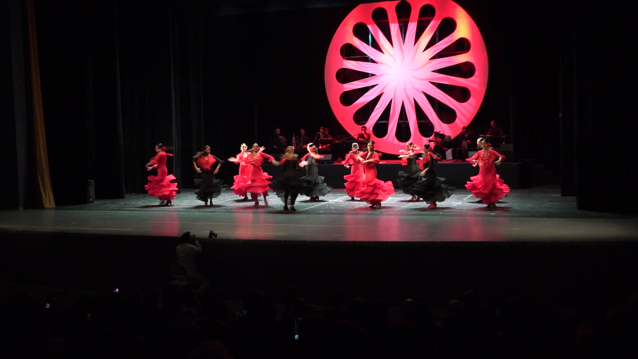 Dancers in red and black costumes on a theater stage with a large glowing red circular prop.