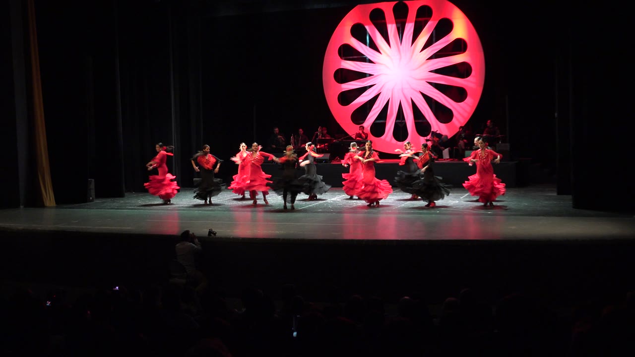 Wide frame from the audience shows the Cabo San Lucas stage performance with dancers arranged across the floor and a glowing red circular prop centered behind them.