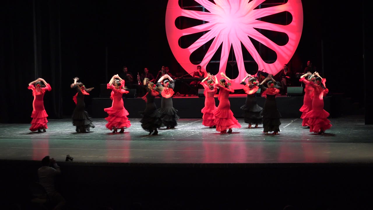 Dancers in red and black dresses perform on a stage with a red fan-shaped backdrop.