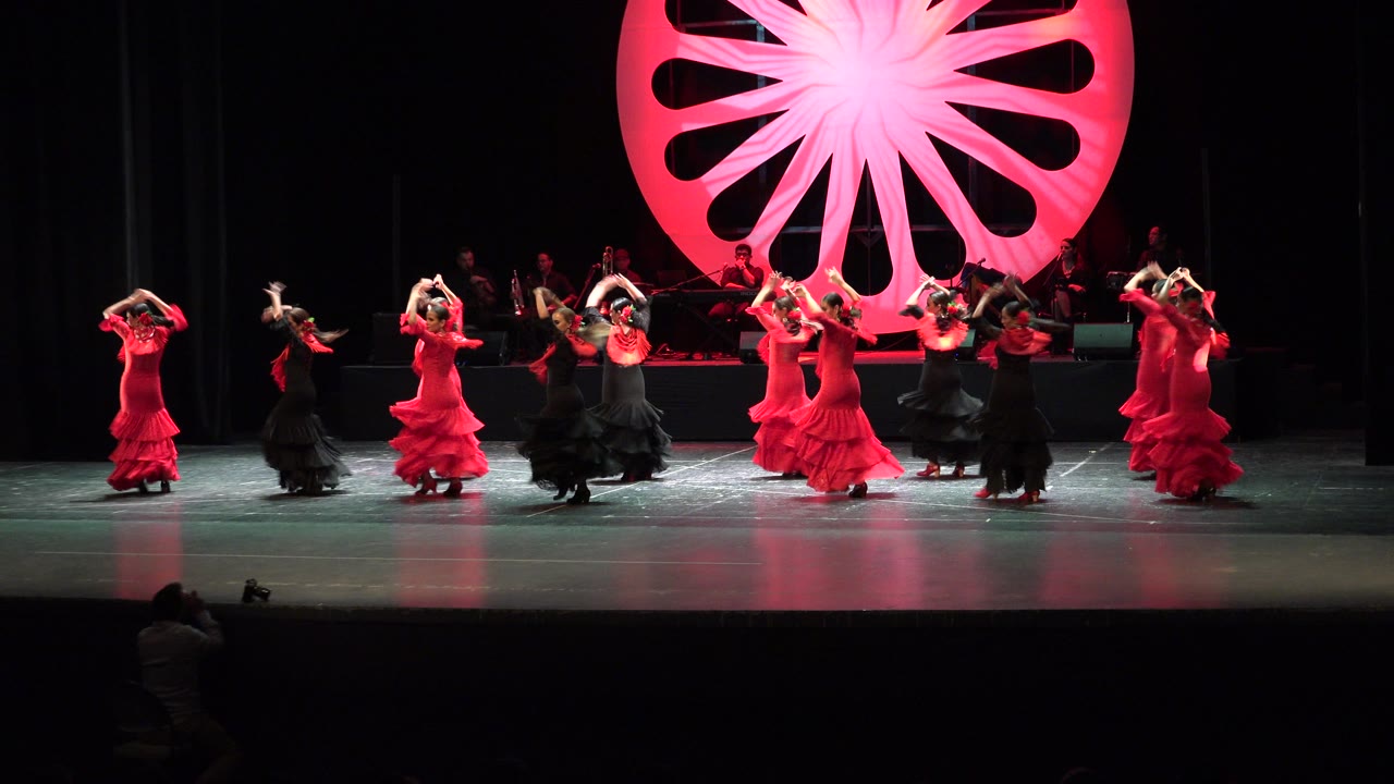 Wide Cabo San Lucas stage frame with the dance group shifting positions, red dresses and black dresses alternating in front of the illuminated circular fan backdrop.