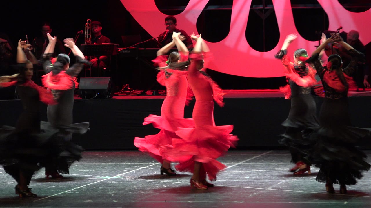 Women in red and black flamenco dresses perform on a dark stage in Cabo San Lucas, Mexico, with musicians visible behind them under red lighting.