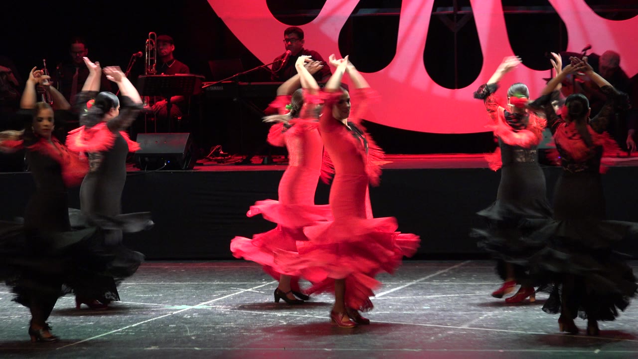 The dancers continue across the red-lit stage in Cabo San Lucas, Mexico, with black dresses in the foreground and a red dress turning near center.