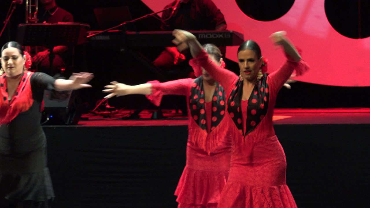 At 0:01 in Cabo San Lucas, Mexico, three women remain near the front of the stage while red sleeves and skirts blur with fast arm and body movement.