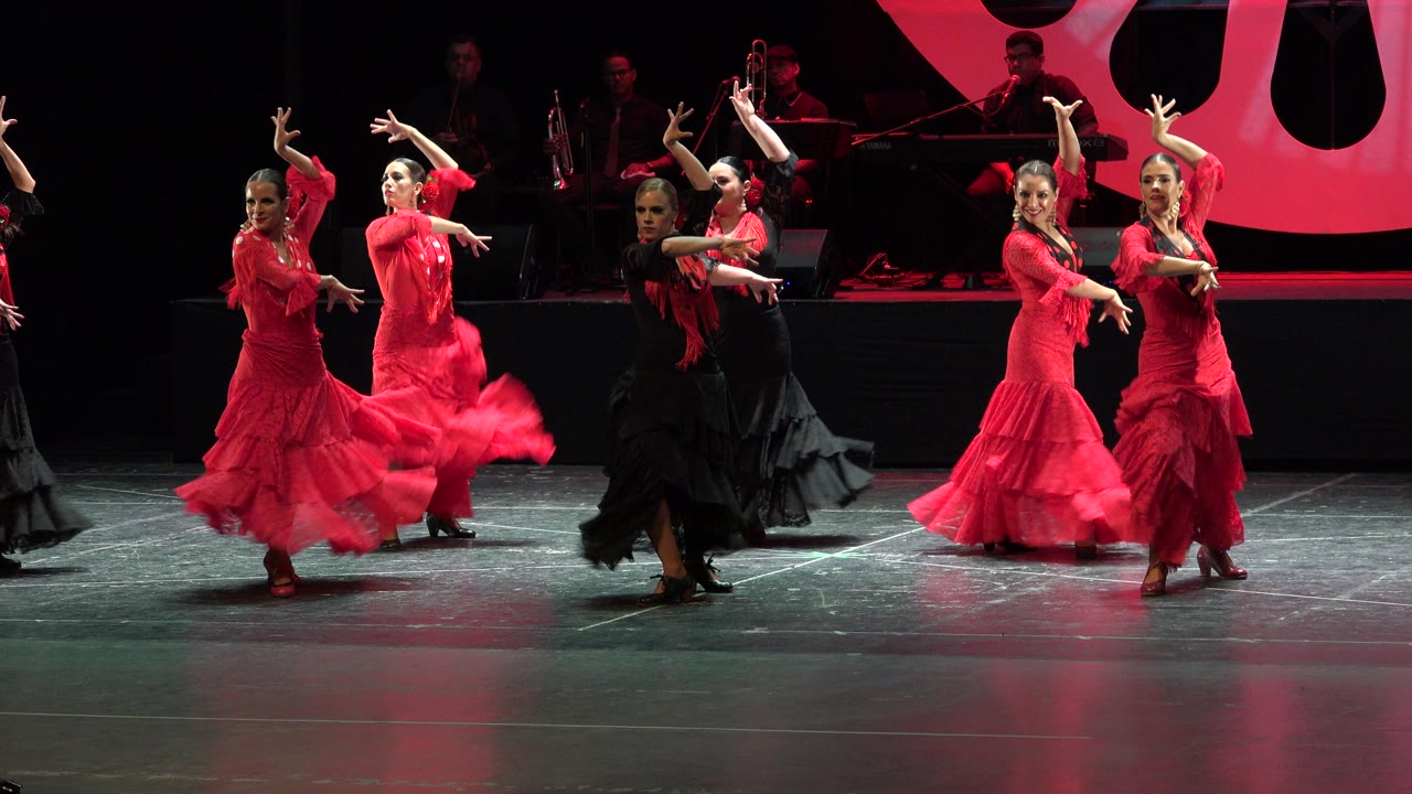 At the opening moment, women in red dresses and one dancer in black perform on a Cabo San Lucas stage with musicians dimly visible behind them.