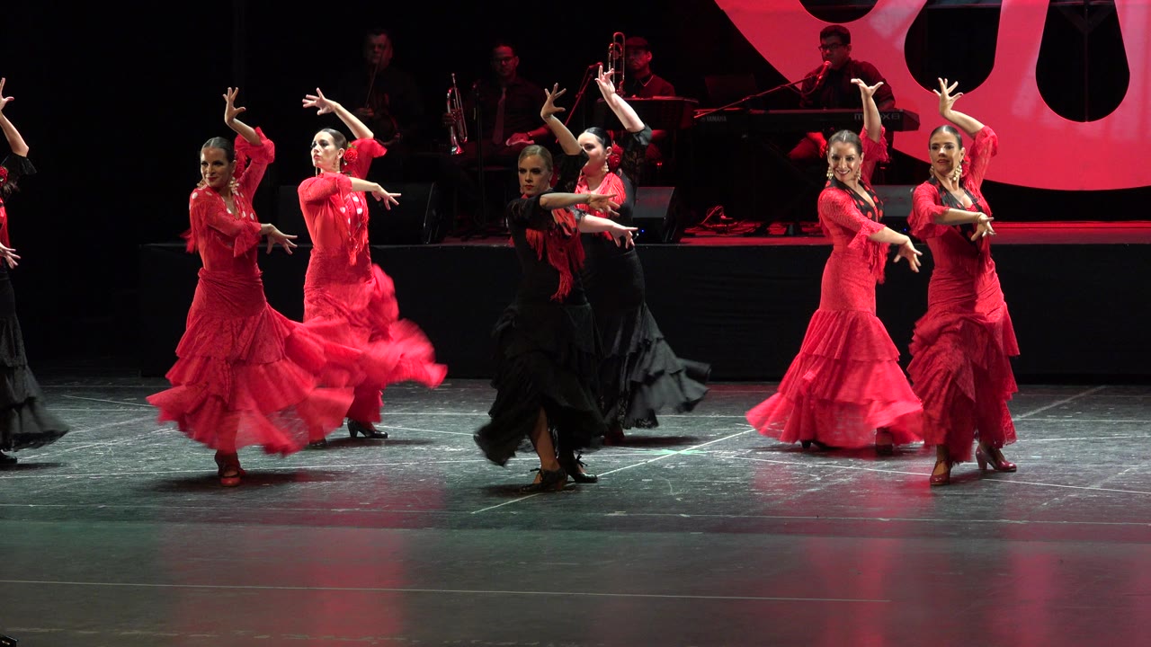 One second in, the Cabo San Lucas dancers hold extended arm positions, with red skirts spread across the lit floor and the band remaining in shadow.