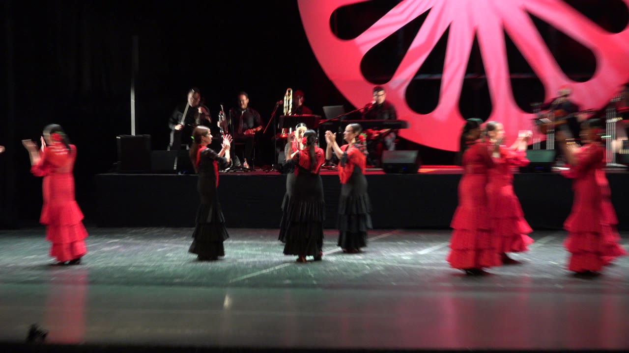 Dancers in red and black costumes perform on a theater stage with musicians behind them.