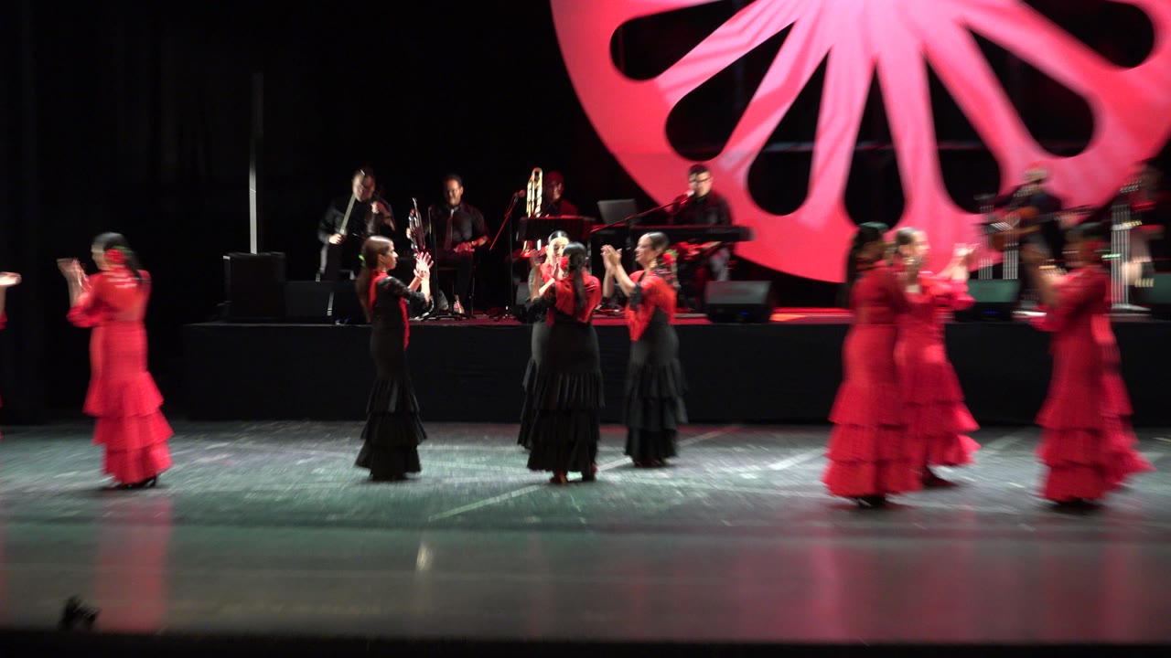 In Cabo San Lucas, Mexico, dancers gather near center stage with hands raised while red-costumed performers blur at the edges and musicians remain on the rear platform.