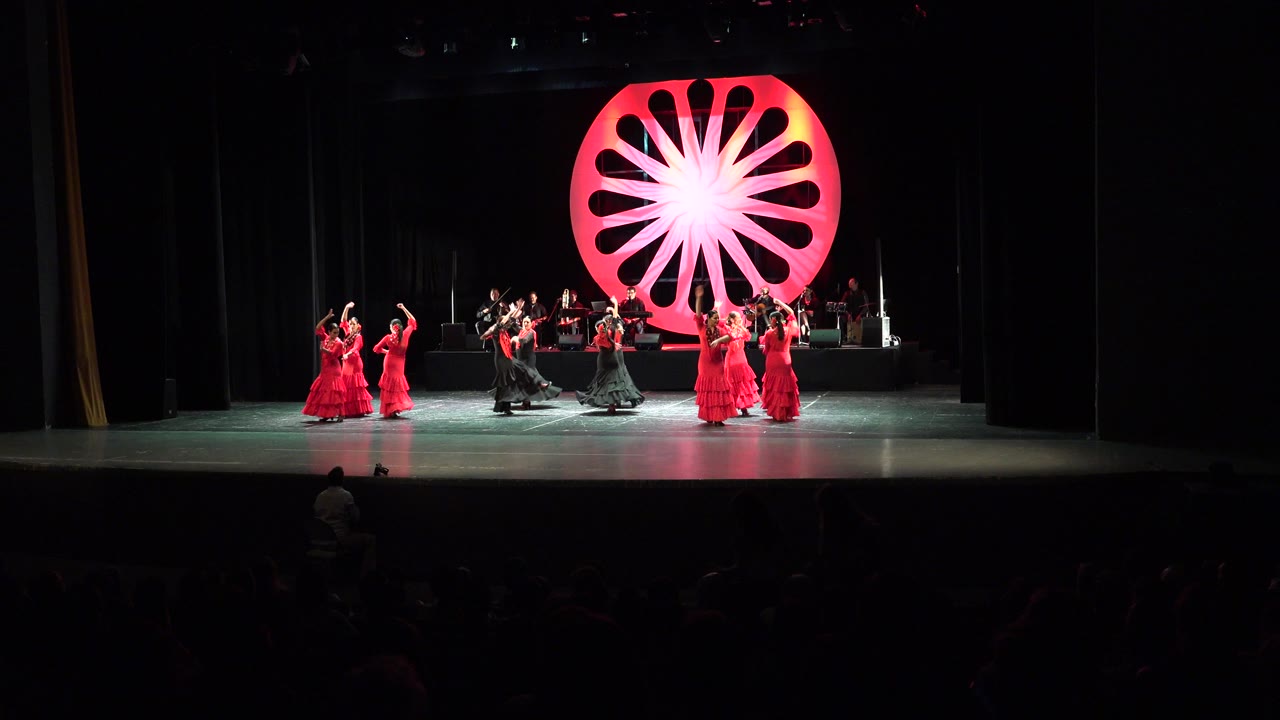 Wide view of a Cabo San Lucas theater stage with red-dressed dancers, black-costumed performers, musicians on a rear riser, and a glowing red circular backdrop.