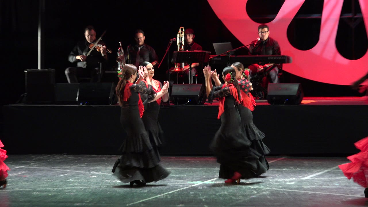 Dancers in black and red costumes perform before seated musicians on a stage.