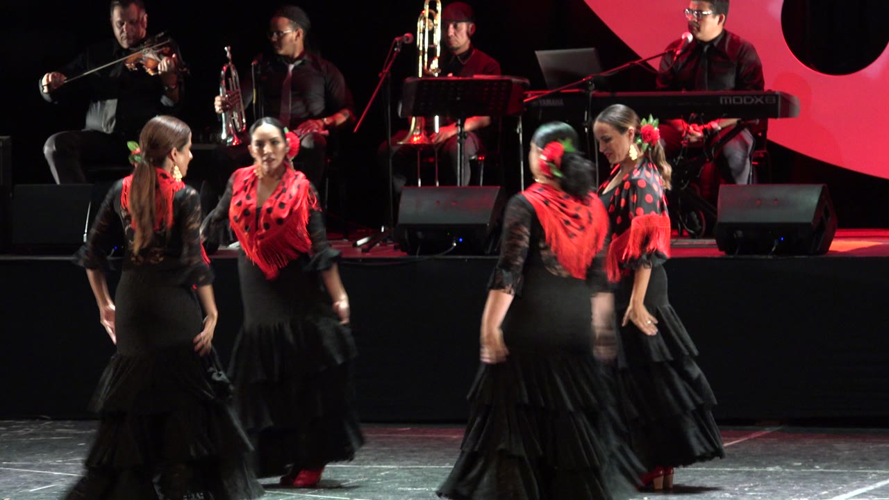 In Cabo San Lucas, Mexico, girls in black dresses with red shawls perform on a stage while musicians play violin, trumpet, trombone, and keyboard behind them.