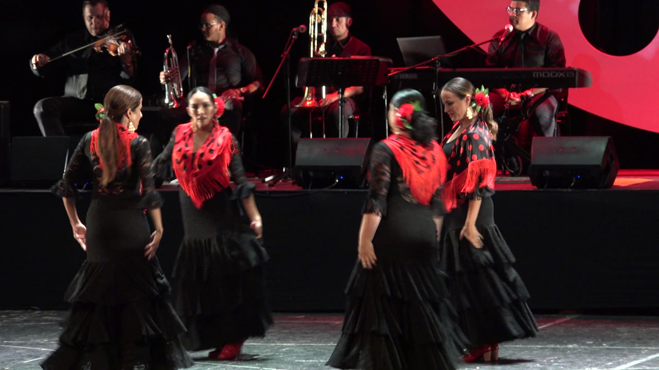 In Cabo San Lucas, Mexico, three girls in black dresses and red shawls hold a formation near the front of the stage with brass and keyboard musicians in the background.