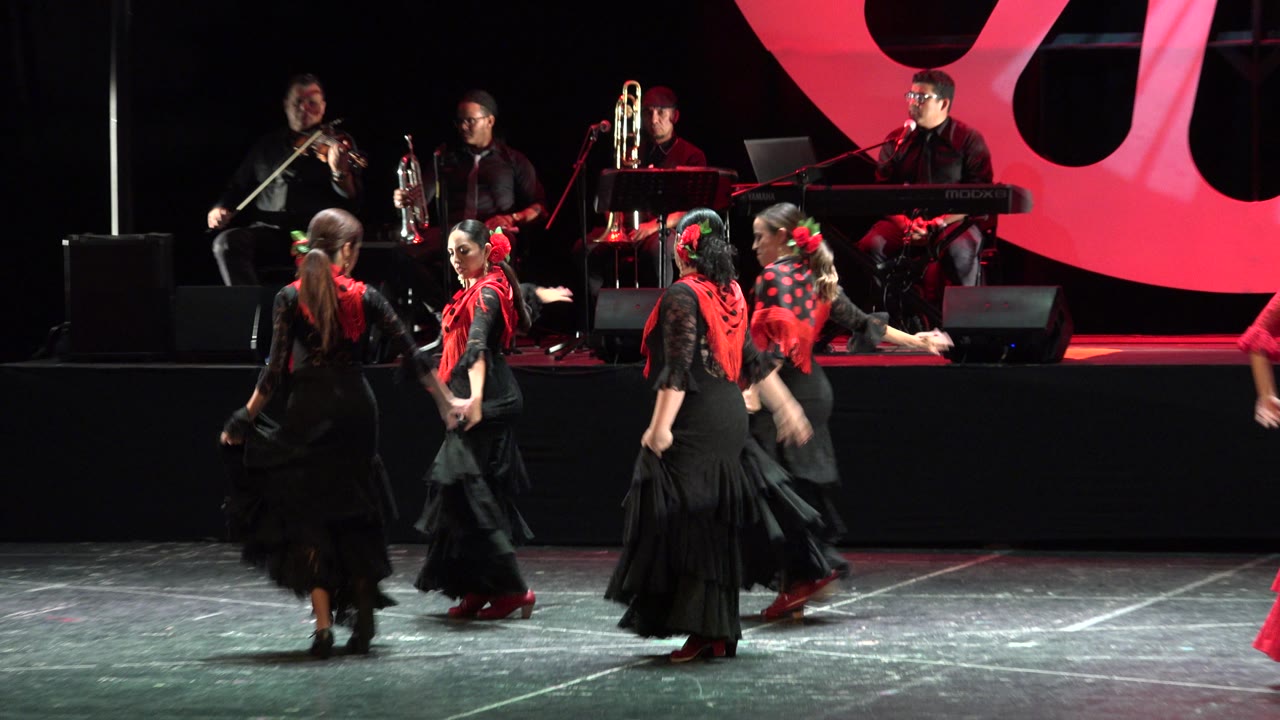 Dancers in black and red move across the stage with the band elevated behind them.