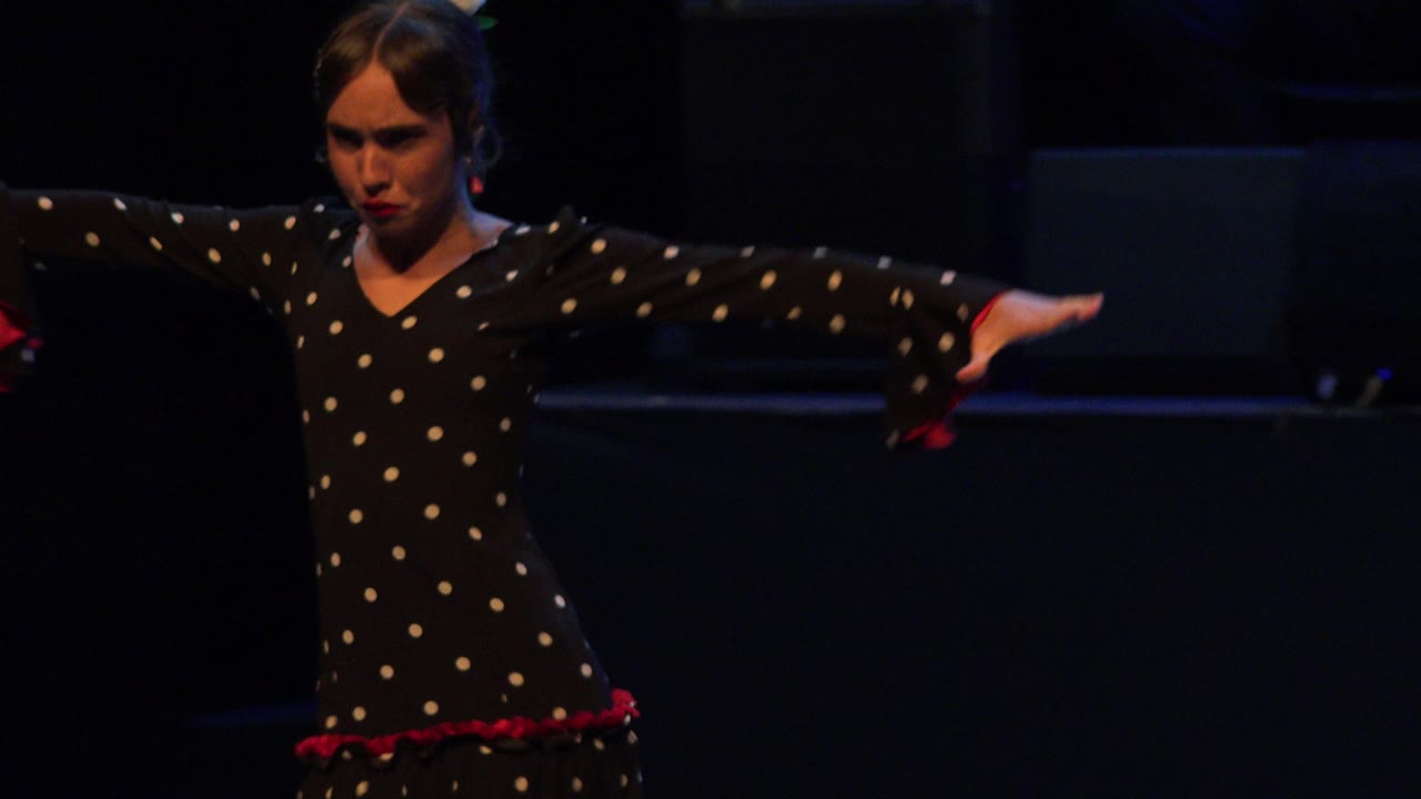 Girl in a black polka-dot dress dancing with arms extended on a dark stage.