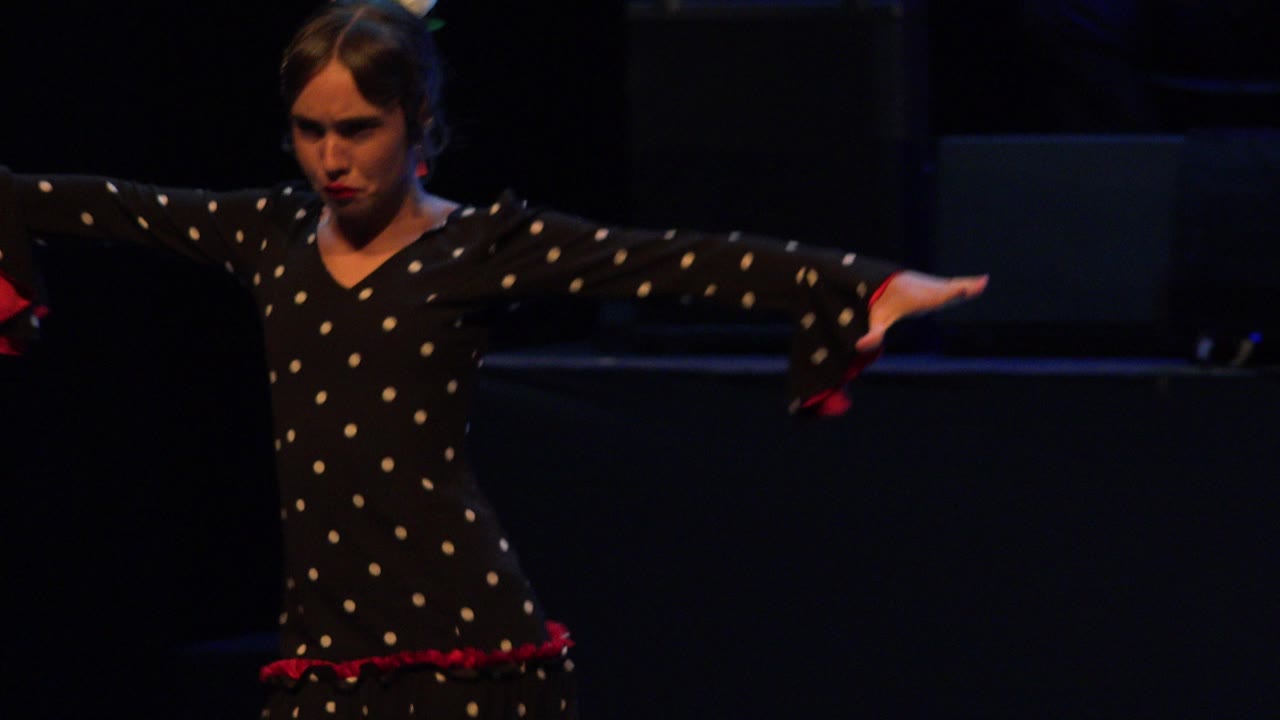 The girl continues the dance with both arms stretched outward, her black polka-dot dress and red cuffs visible against the dark Cabo San Lucas stage.