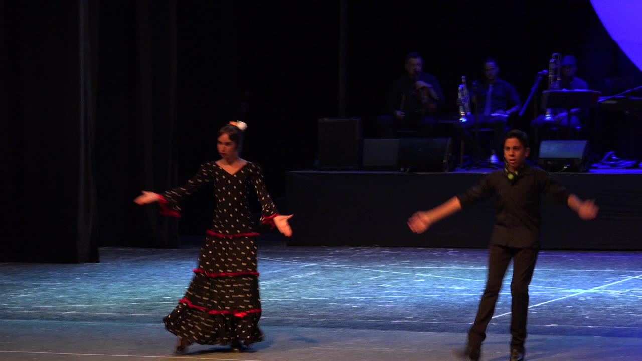 The Cabo San Lucas stage performance continues with the woman gesturing outward and the male dancer moving his arms beside her under blue-purple stage light.