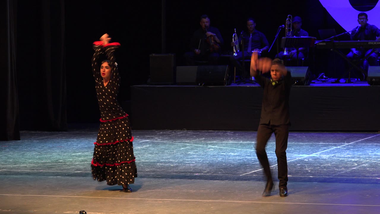 Female dancer holds both arms overhead as male dancer moves beside her.