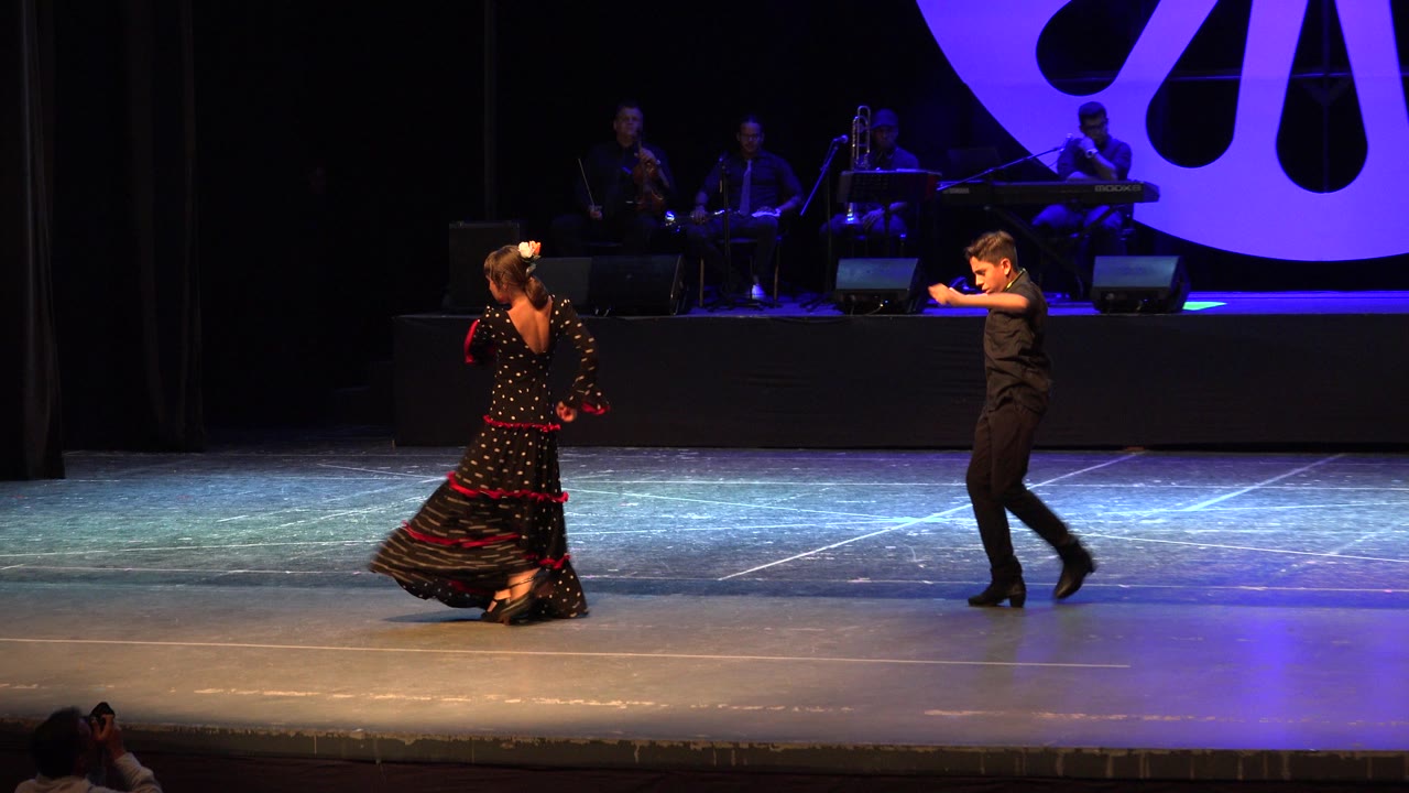 Two young dancers perform on a Cabo San Lucas theater stage, with the girl in a black and red dress facing a boy in black as musicians sit in blue-purple light behind them.